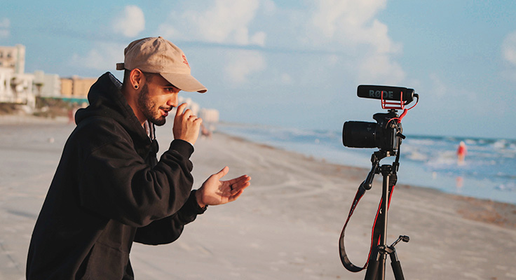 Mann som filmar seg sjølv med kamera på ein tripod på ei strand